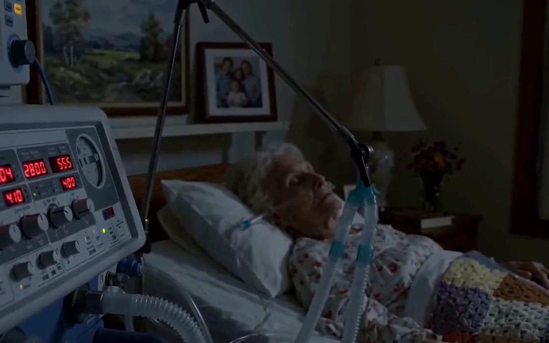 An elderly patient asleep in a hospital bed beside a ventilator with red digital readouts.