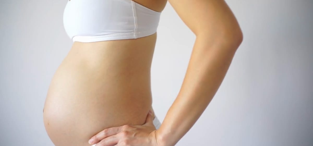 Side view of a pregnant woman in a white top, one hand resting on her hip.