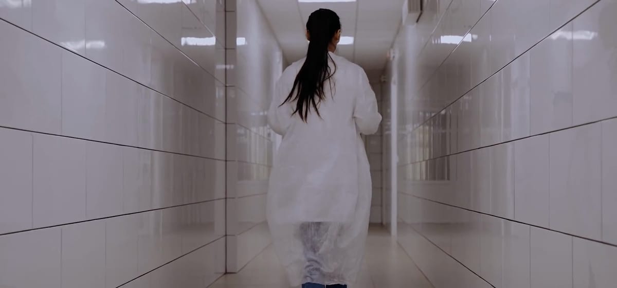 A woman in a white lab coat walks away down a long, bright hospital corridor.
