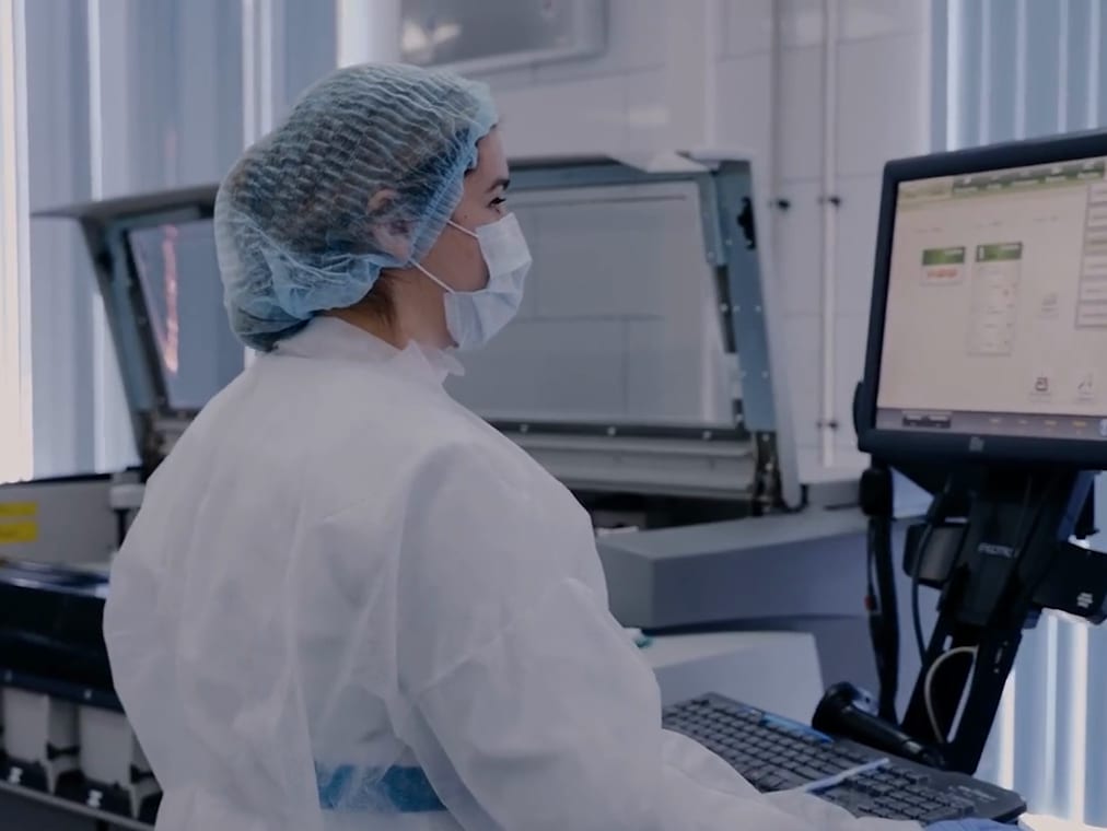 A technician in a hairnet and mask working at an analyser's touchscreen in a clean room.