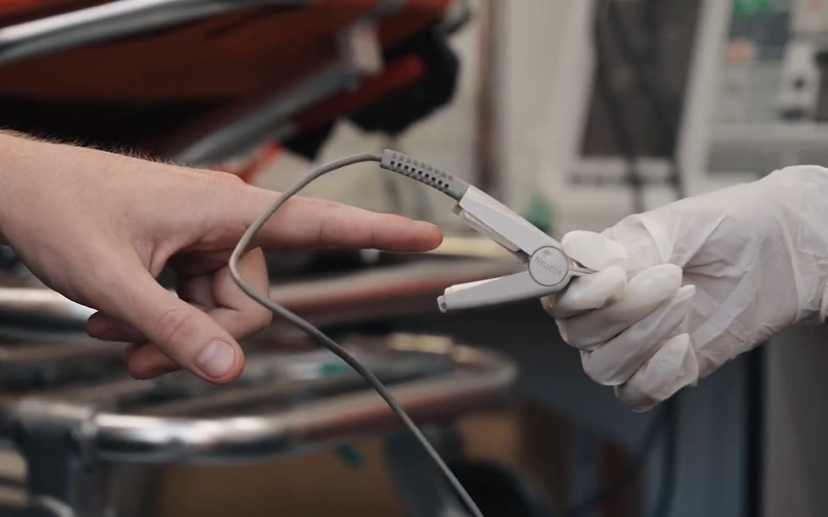 A gloved hand clips a pulse oximeter sensor onto a patient's finger beside a hospital bed.